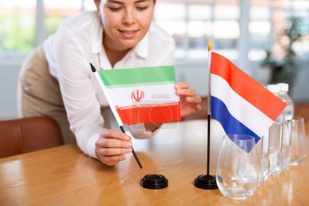 Unrecognizable woman preparing room for international negotiations and communication discussions of leaders. Lady sets miniatures flags of Netherlands and Iran on table. Unfocused shot
