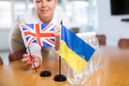 Closeup of national flags of Ukraine and United Kingdom in hands of female office secretary preparing table in meeting room for international negotiations. Concept of diplomatic relations
