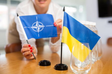 Flaggen der NATO und der Ukraine in den Händen einer Bürokoordinatorin, die den Sitzungssaal für strategische Verhandlungen über Sicherheit und Stärkung der Zusammenarbeit vorbereitet