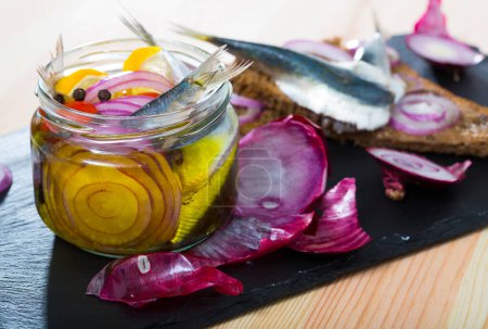 Jar of homemade marinated sprats with onion, beetroot, assorted peppers, vinegar, lemon, olive oil, sea salt. Served with rye bread on slate plate