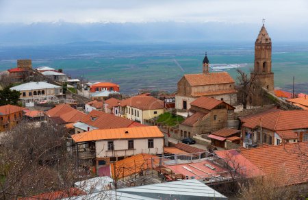 Scenic spring view of tiled roofs of houses and Church of St. George with bell tower in Georgian township of Sighnaghi on background of Alazani valley and snow-capped Greater Caucasus ridge on horizon