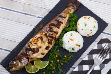 Köstlicher gebackener Forellenfisch auf schwarzem Brett mit Garnitur aus weißem Reis, gegrillten Zwiebelringen, Gemüse, Erbsen und Zitronenscheiben