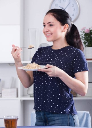 Young smiling girl standing at table and eating cereal for breakfast