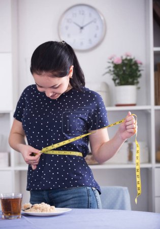 Smiling young woman measuring waist with tape near table with breakfast