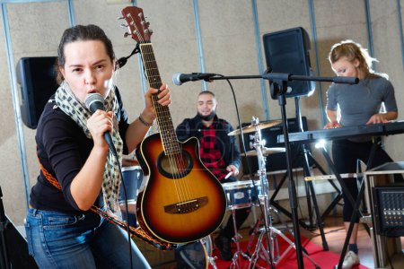 Attractive satisfied pleasant friendly female soloist playing guitar and singing with her music band in sound studio
