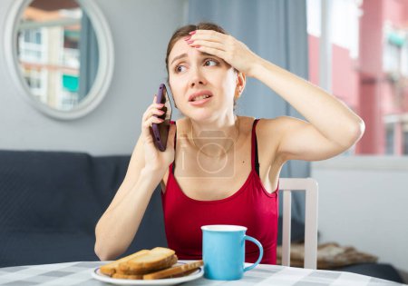 Mujer disgustada hablando por teléfono móvil mientras está sentada en la mesa de la cena en casa