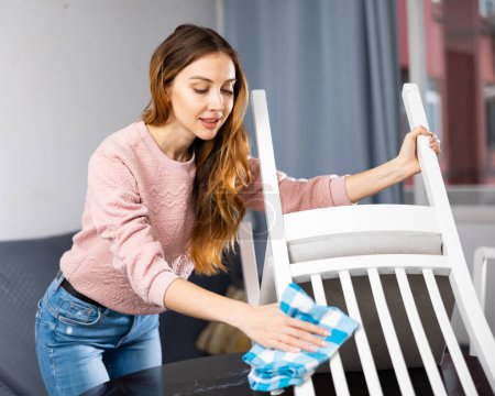 Hardworking young woman wipes a chair with a rag, tidying up the apartment