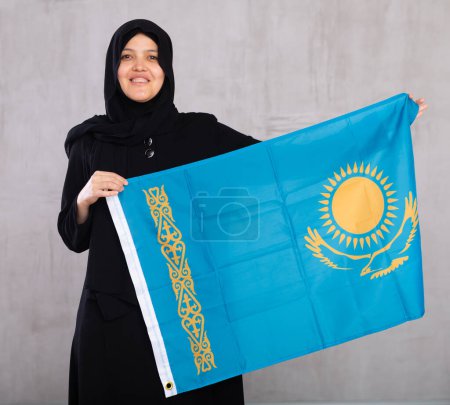 Mujer musulmana joven en hiyab negro sostiene la bandera desplegada de Kazajstán.Studio tiro, fondo gris