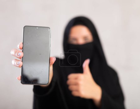 chica musulmana joven con su cara cubierta por burka mantenga en la mano el uso de cerca de teléfono celular móvil con área de espacio de trabajo de pantalla en blanco