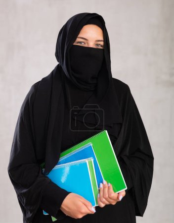 Mädchen mit Gesicht, das mit Burka bedeckt ist, hält viele dicke Notizbücher in der Hand. ausländischer muslimischer Student mit einem Stapel Zettel. Nahaufnahme, grauer Hintergrund