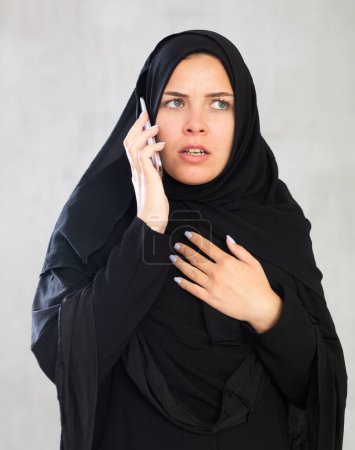 mujer musulmana de piel clara, desalentada en hiyab negro está hablando por teléfono móvil. Calma El estado musulmán femenino está hablando por teléfono. Primer plano sobre fondo gris