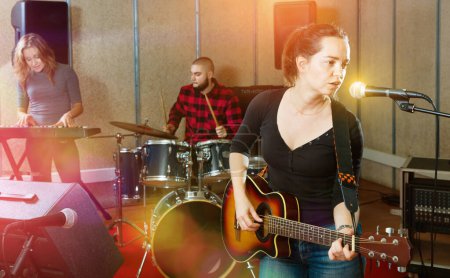 Attractive positive glad pleasant female soloist playing guitar and singing with her music band in sound studio