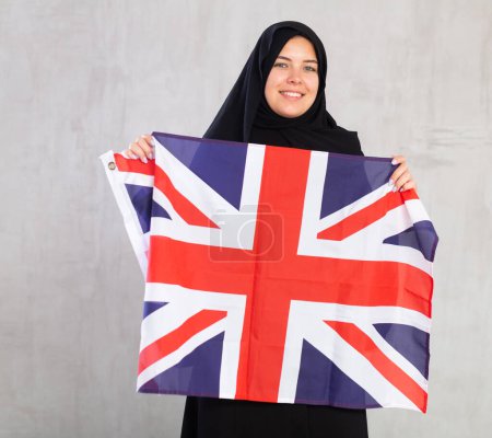 smiling Muslim woman in traditional black hijab holds flag of Great Britain. Portrait of female Muslim with British flag on gray background