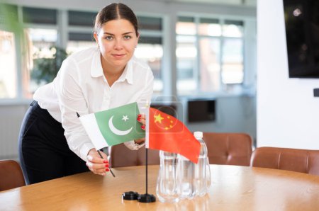 Assistant girl prepares office for international negotiations and meetings of leaders. Lady sets miniatures flags of China and Pakistan on table. Close-up