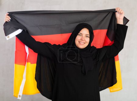 sonriente mujer musulmana en hiyab negro tradicional sostiene la bandera de Alemania. Retrato de mujer musulmana con bandera alemana sobre fondo gris