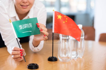 Unrecognizable woman preparing room for international negotiations and communication discussions of leaders. Lady sets miniatures flags of China and Saudi Arabia on table. Unfocused shot