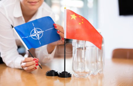 Unrecognizable woman preparing room for international negotiations and communication discussions of leaders. Lady sets miniatures flags of China and NATO on table. Unfocused shot