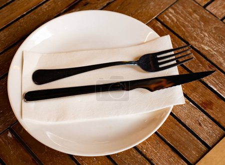 In refreshment bar on wooden table, round plate with cutlery, knife and fork complemented by transparent glass for drinks. Restaurant serving option, folded napkin covers dinner plate.