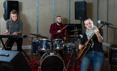 Portrait of active excited girl rock singer with guitar during rehearsal with male drummer and female keyboardist in studio