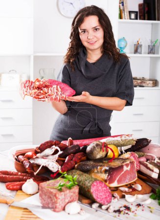 Positive young woman costs near table with smoked products and sausage indoors