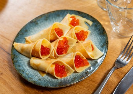There are envelope-shaped pancakes filled with red caviar on plate. Delicacy, snack, hearty dish of national Russian cuisine