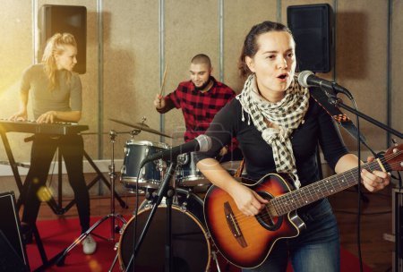 Attractive happy cheerful positive smiling female soloist playing guitar and singing with her music band in sound studio