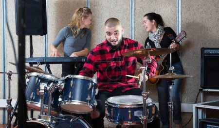 Group of young musicians with expressive male drummer rehearsing in rehearsal room