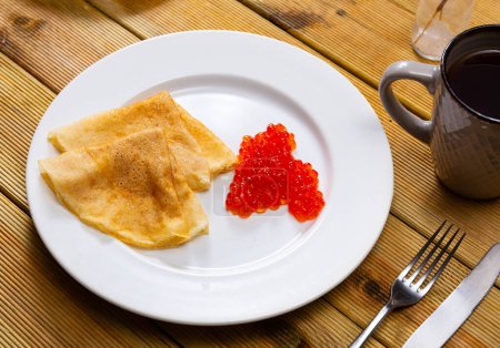 On plate are several rolled delicate pancakes with ruddy edge and spoonful red salmon caviar. Hearty snack is complemented by cup of fragrant coffee, tea