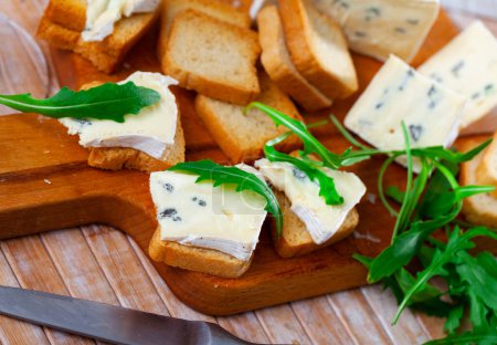 On cutting board there is slicing of blue cheese and rusk, complemented decorated with fresh arugula leaves. Large board with appetizer stands on wooden table next to glass