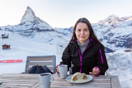 Woman enjoying fragrant mulled wine and slice of apple strudel topped with vanilla sauce in winter day while traveling in Swiss Alps, sitting at table on background of Matterhorn mountain..