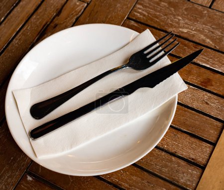 In refreshment bar on wooden table, round plate with cutlery, knife and fork complemented by transparent glass for drinks. Restaurant serving option, folded napkin covers dinner plate.