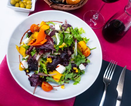 Colorful salad of fresh lettuce and arugula leaves with ripe tomatoes, chopped mango, soft cheese and balsamic sauce.