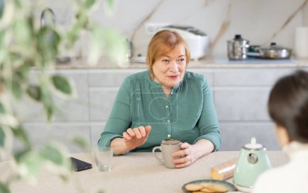 Daheim in der Küche am Tisch sitzt eine ältere lächelnde Frau und hat eine interaktive Beziehung zu einem Freund und trinkt Kaffee oder Tee. Konzept lebendige Beziehung und persönliche Gespräche
