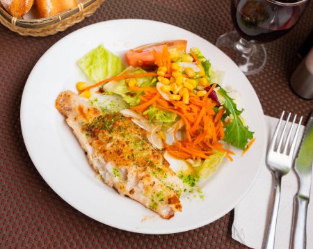 Appetizing deep fried fish fillet, chopped parsley and garlic in olive oil served with fresh vegetable salad on plate.
