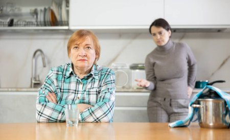 Young girl makes claims her mother emotionally.Sad mature woman with soup in pan on table listens to reproaches of daughter sitting on kitchen at home. Misunderstanding relationship