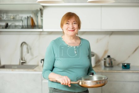 Portrait d'une femme mature positive et confiante de bonne humeur, debout dans la cuisine à la maison et tenant une poêle .in ses mains