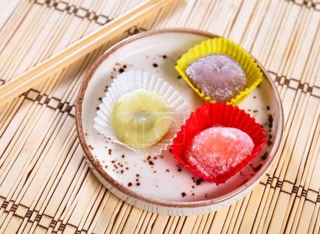 Colorful mochi desserts with various delicate flavors in yellow, white, and red paper liners, arranged on ceramic plate with chopsticks over bamboo mat. Sweet delicacy in Japanese style