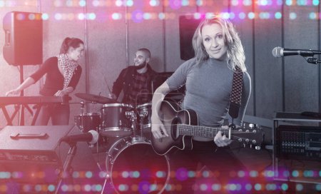Portrait of excited young blonde girl rock singer with guitar during rehearsal with male drummer and female keyboardist in studio