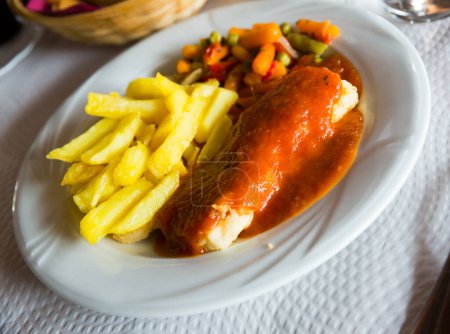 Filete de bacalao apetitoso cocido en salsa de tomate servido con guarnición vegetal..