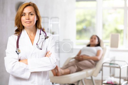 Médecin d'âge moyen positif d'apparence européenne debout à l'intérieur du bureau médical sur fond de femme menteuse