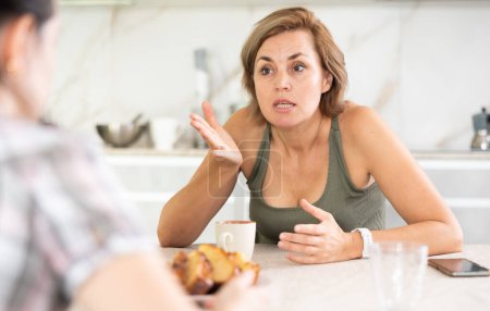 Mujer de mediana edad trastornada hablando tristemente con su colaboradora mientras bebe té en la cocina