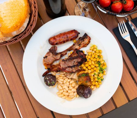 Grilled cold cuts with legumes are on plate. Grilled lamb ribs, sausage, piece of black pudding are complemented with stewed beans, green peas. There is bowl of cherry tomatoes in background