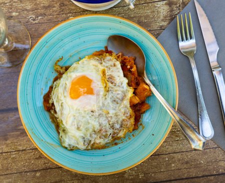 Delightful Spanish cuisine dish of cod in rich tomato sauce with vegetables and garlic topped with sunny side up egg, presented on rustic wooden table with glass of beer