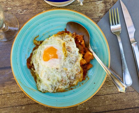 Delightful Spanish cuisine dish of cod in rich tomato sauce with vegetables and garlic topped with sunny side up egg, presented on rustic wooden table with glass of beer