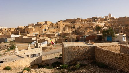 Dilapidated stone houses stacked on hillside in ancient Berber settlement of Tamezret with mosque minaret rising above under hot Tunisian sun