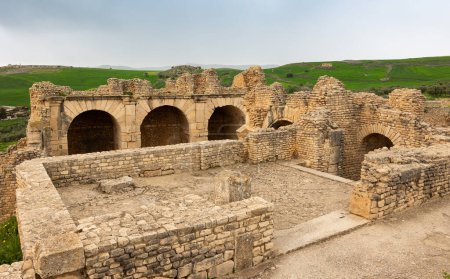 Blick auf Ruinen des antiken römischen Nymphäums in der tunesischen Siedlung Dougga mit gut erhaltenen Bögen und Mauerresten an bewölkten Frühlingstagen