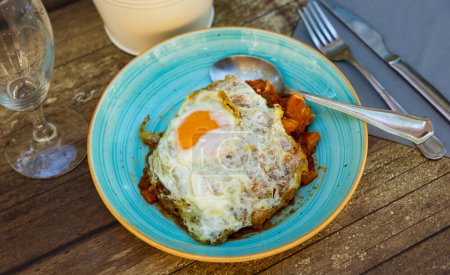 Rich cod stew with mix of vegetables in garlic and tomato salsa, topped with fried egg, served on rustic wooden table. Traditional recipe from Basque Country cuisine