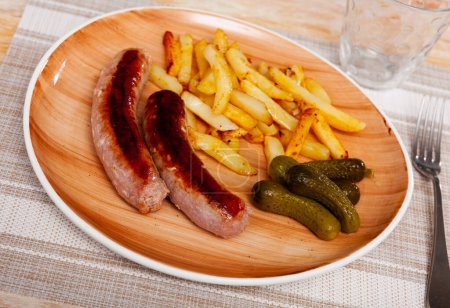 Tasty hearty lunch of roasted meat sausages with french fried potatoes and pickled cucumbers