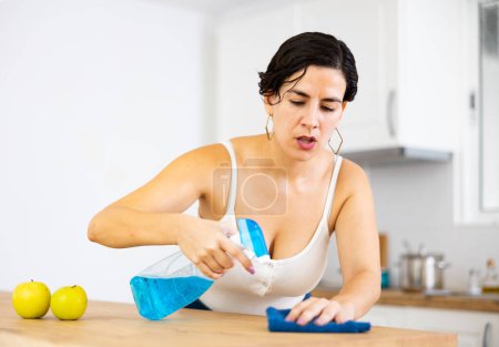 Porträt einer fokussierten jungen hispanischen Frau, die Tisch mit Tuch und Spray in der Küche reinigt. Konzept für alltägliche Hausarbeit