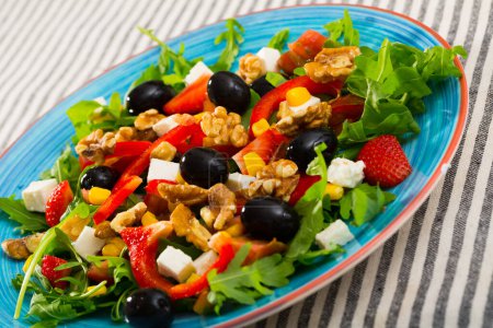 Vitamin salad of arugula with tomatoes, red bell pepper, feta cheese, strawberry and walnuts in plate on striped textile background